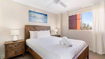 A hotel room with a large bed, wooden headboard, two bedside tables with lamps, a ceiling fan, a window with curtains, and a painting on the wall.