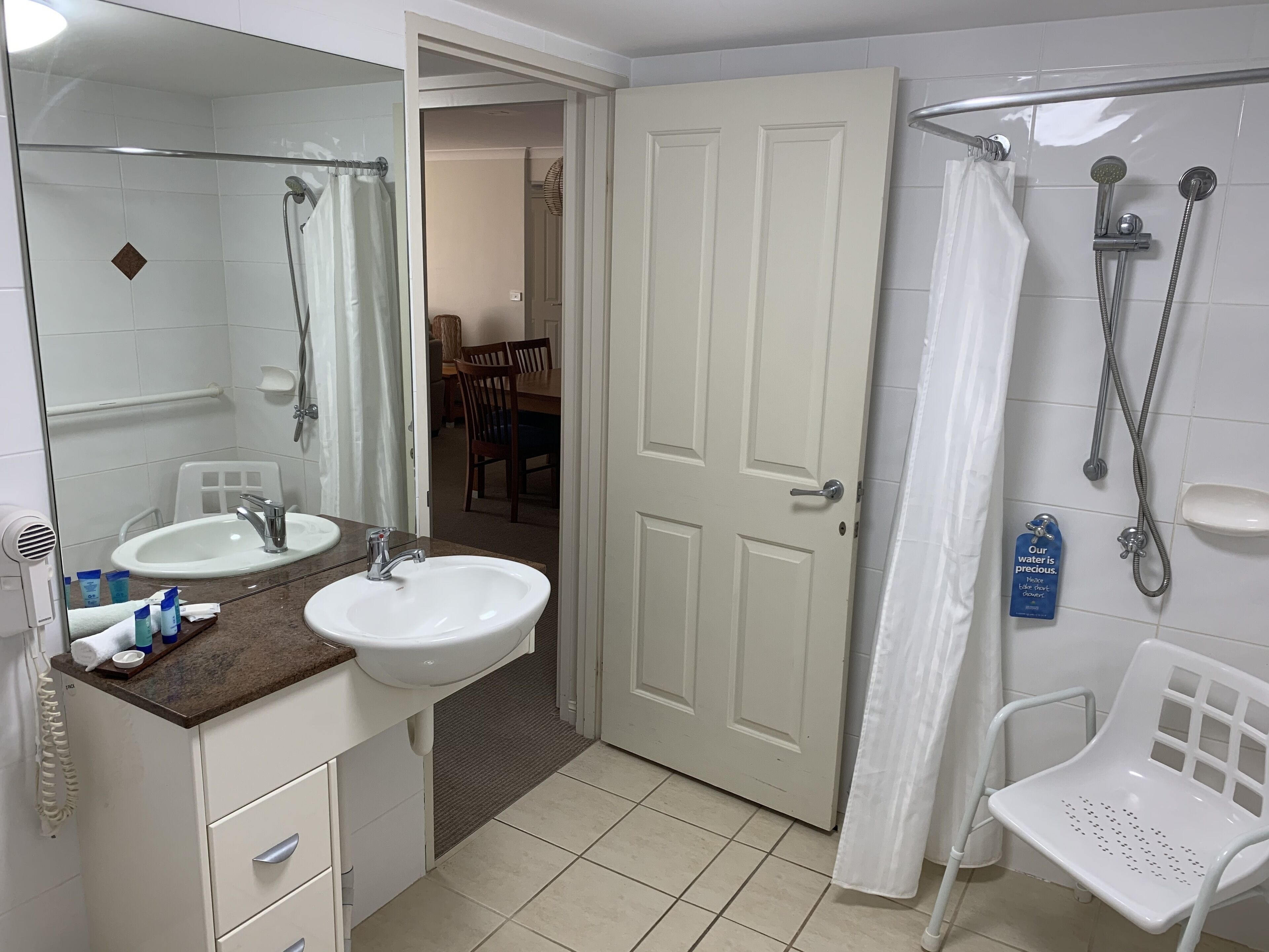 Ein Badezimmer mit zwei Waschbecken, einer Dusche und einer Toilette. Über den Waschbecken hängt ein Spiegel, und ein Handtuchhalter ist zu sehen.