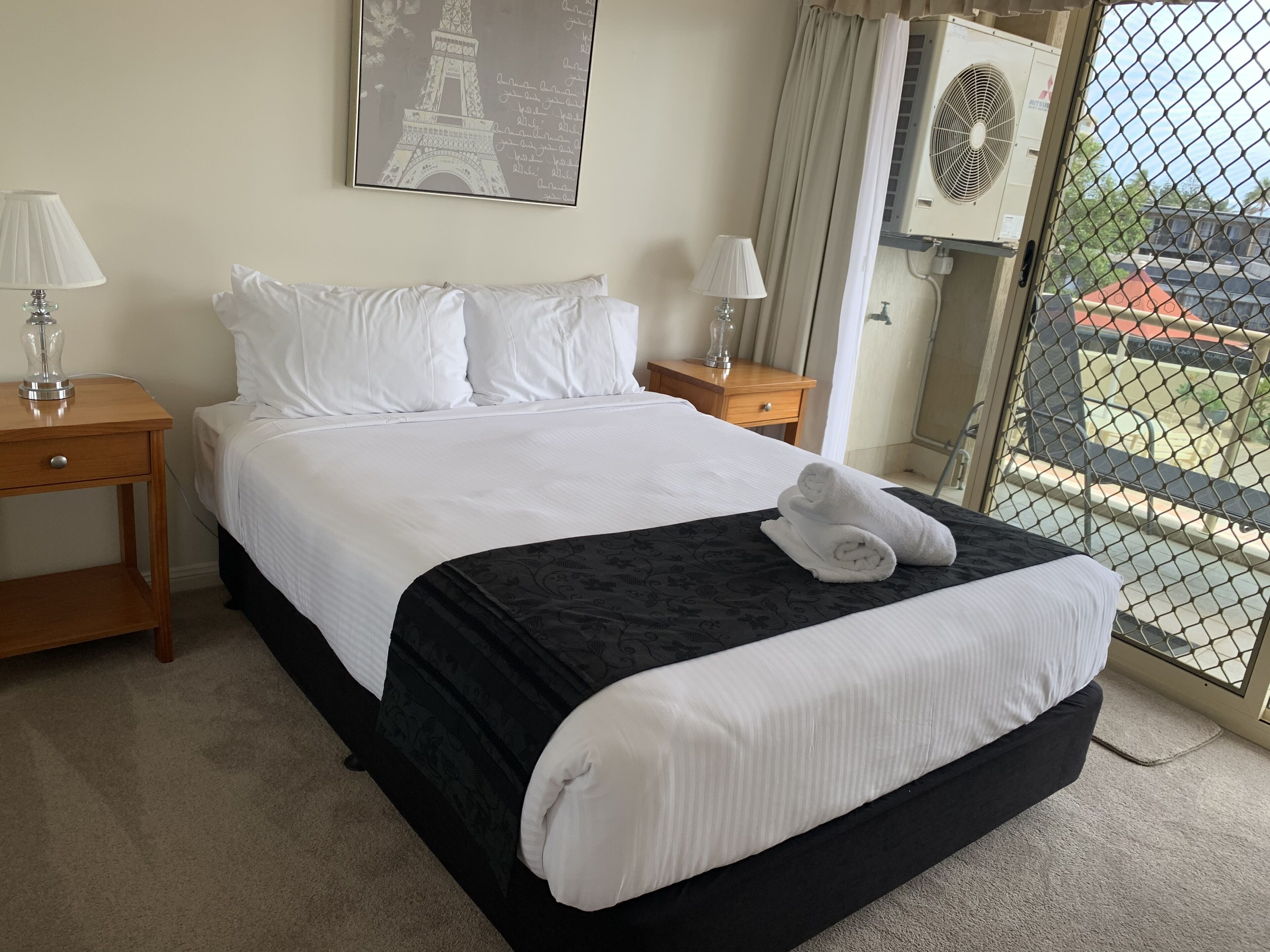 A hotel room with a bed, two bedside tables, a nightstand, a window with a view, and an air conditioning unit.