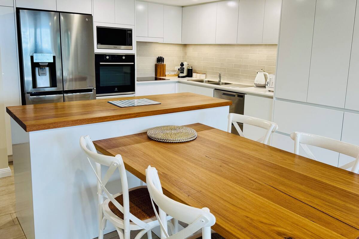 Cocina moderna con armarios blancos, un refrigerador de acero inoxidable, horno empotrado, microondas y lavavajillas. Hay una mesa de madera con dos sillas blancas.