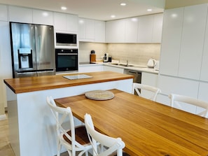 A modern kitchen with white cabinetry, a stainless steel refrigerator, a built-in oven, a microwave, and a dishwasher. There is a wooden dining table with two white chairs.
