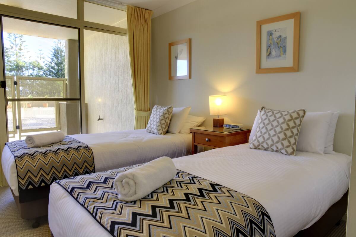 Habitación de hotel con dos camas, una mesita de noche, una lámpara y una ventana con cortinas.