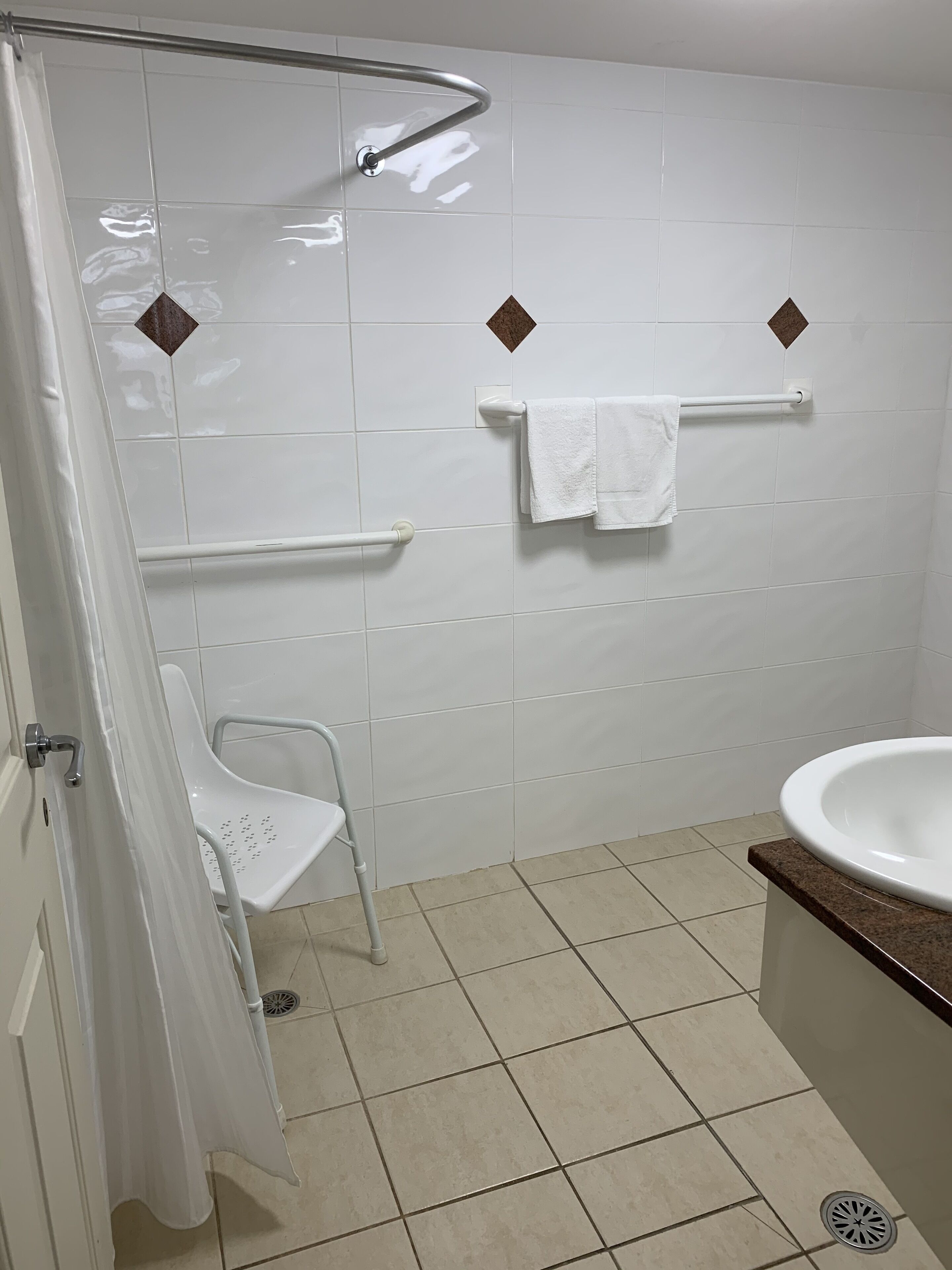 A bathroom with white tiles, a shower with a grab bar, a white toilet with a safety bar, and a sink with a marble countertop.