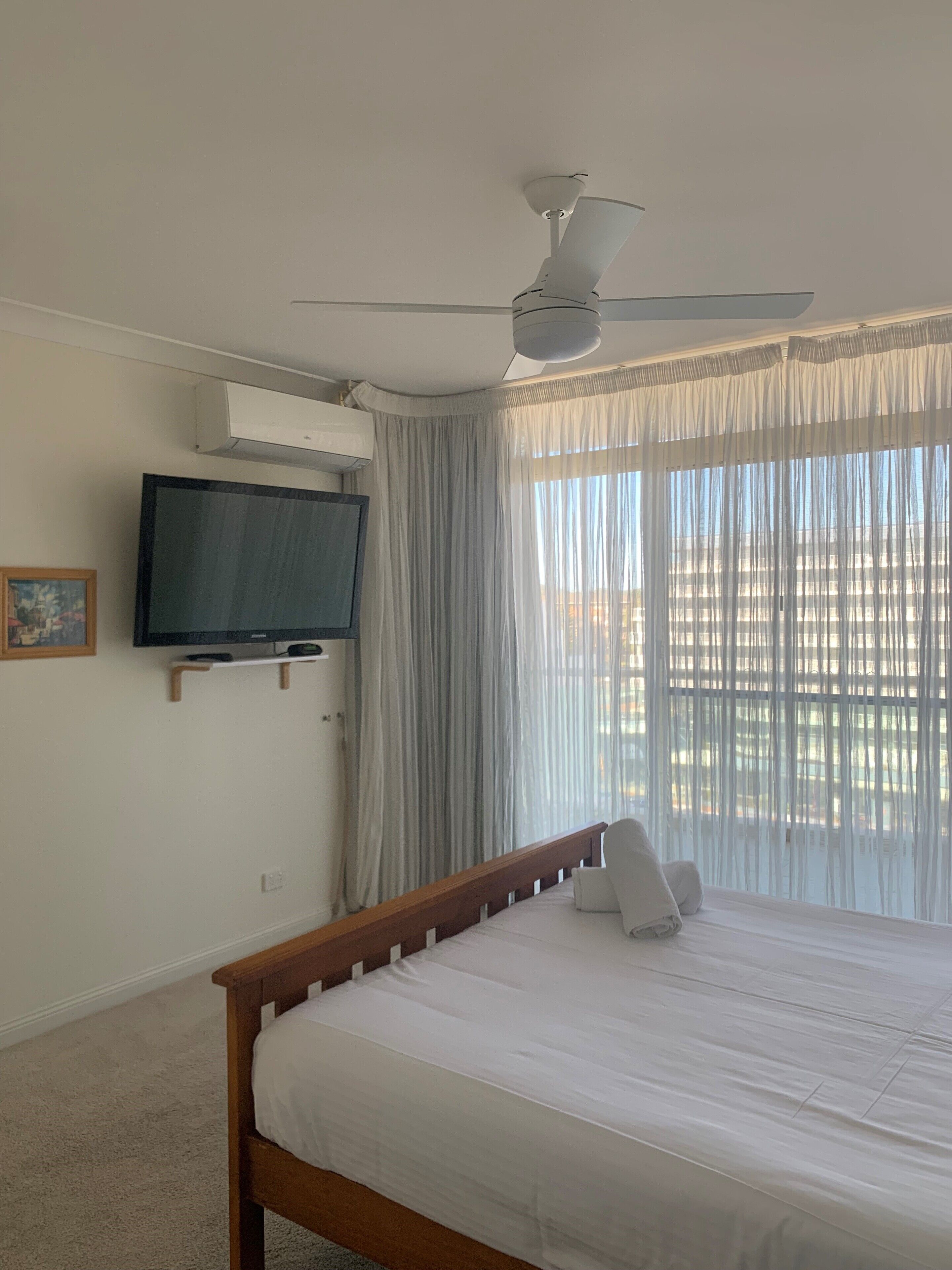 A bedroom with a bed, a ceiling fan, a TV mounted on the wall, and sheer curtains.