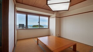 Um quarto com piso de tatame, uma mesa de madeira e uma grande janela com vista para o mar e uma ilha distante.