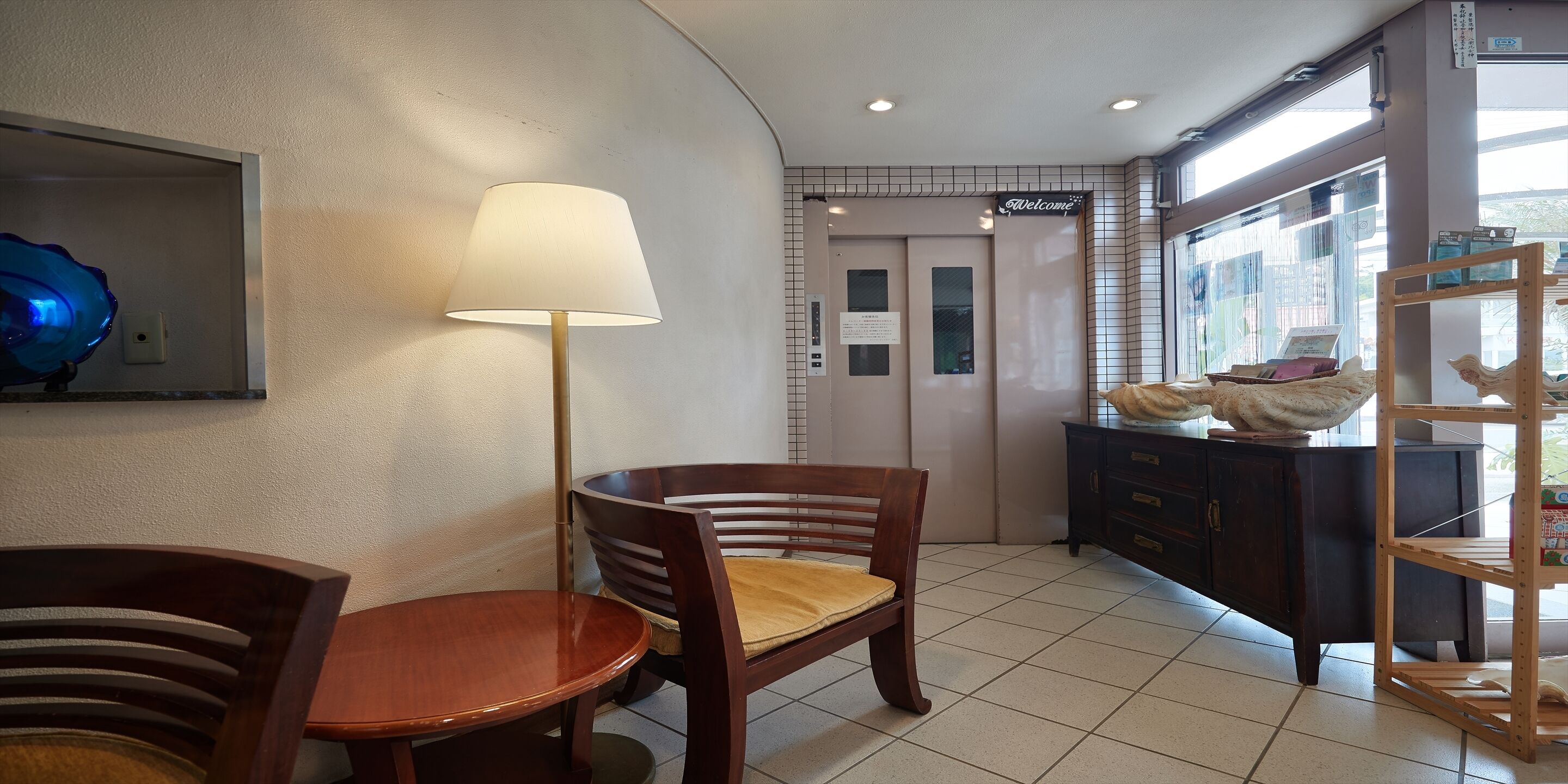 Um hall de hotel com uma cadeira e mesa de madeira, uma luminária, um balcão de recepção e vista pela janela.