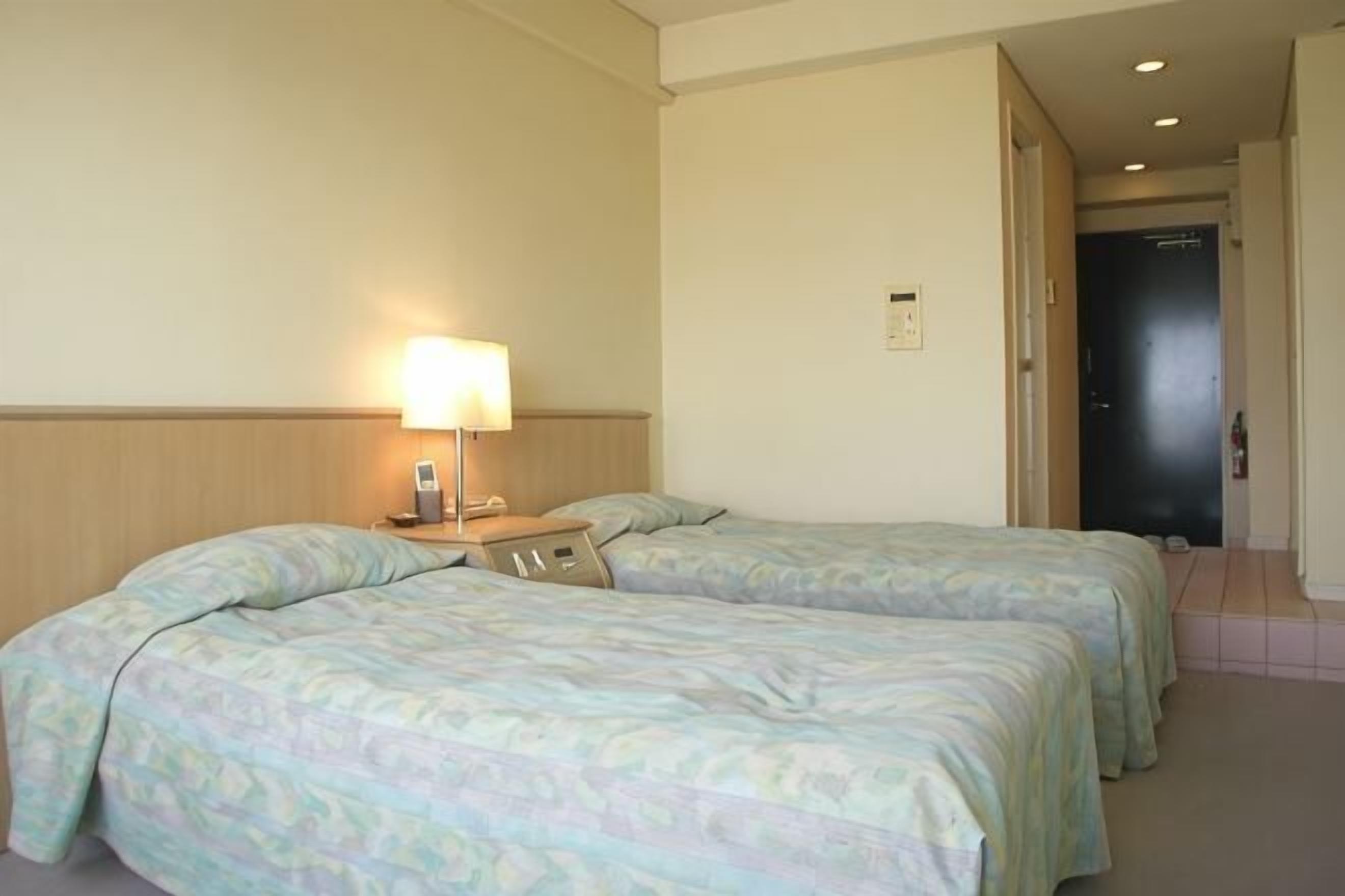 Una habitación de hotel con dos camas, una mesita de noche con lámpara, y una puerta que da a otra habitación.
