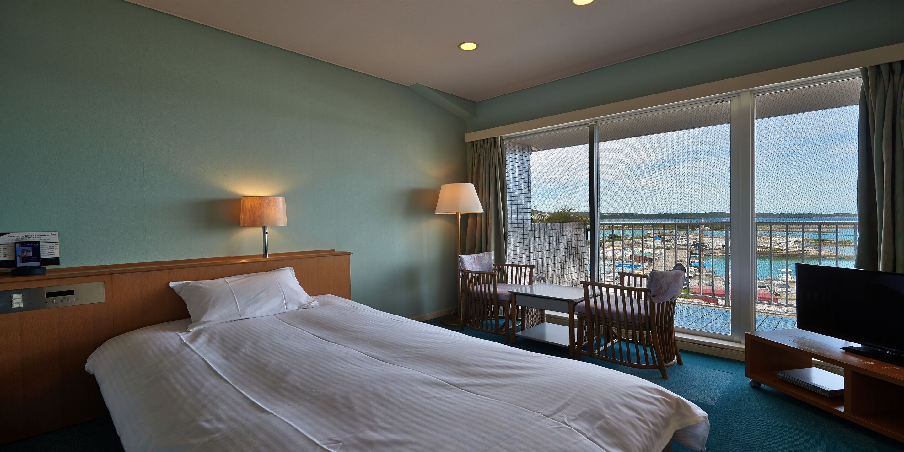 A hotel room with a bed, a desk, a chair, a TV, and a large window with a view of a beach and pool.