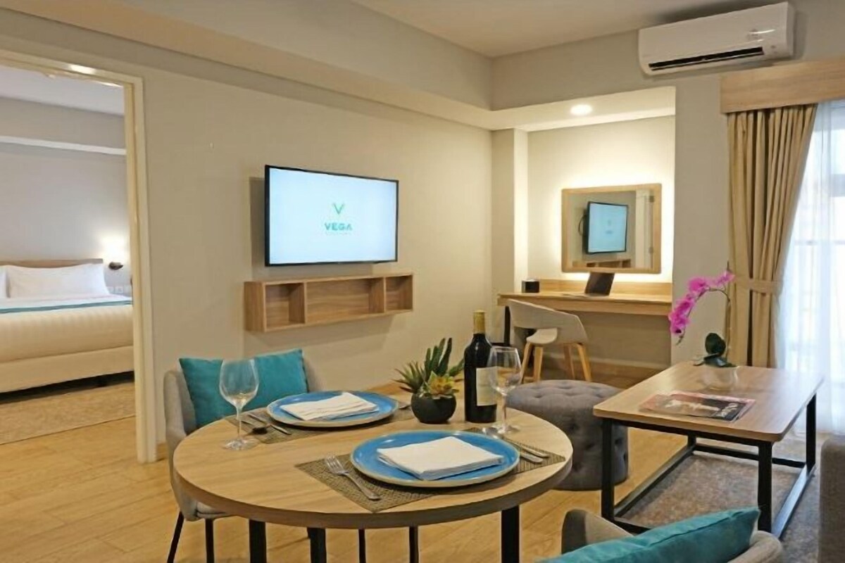 A hotel room with a bed, a TV, a desk, a chair, a table with a bottle, glasses, and a plant.