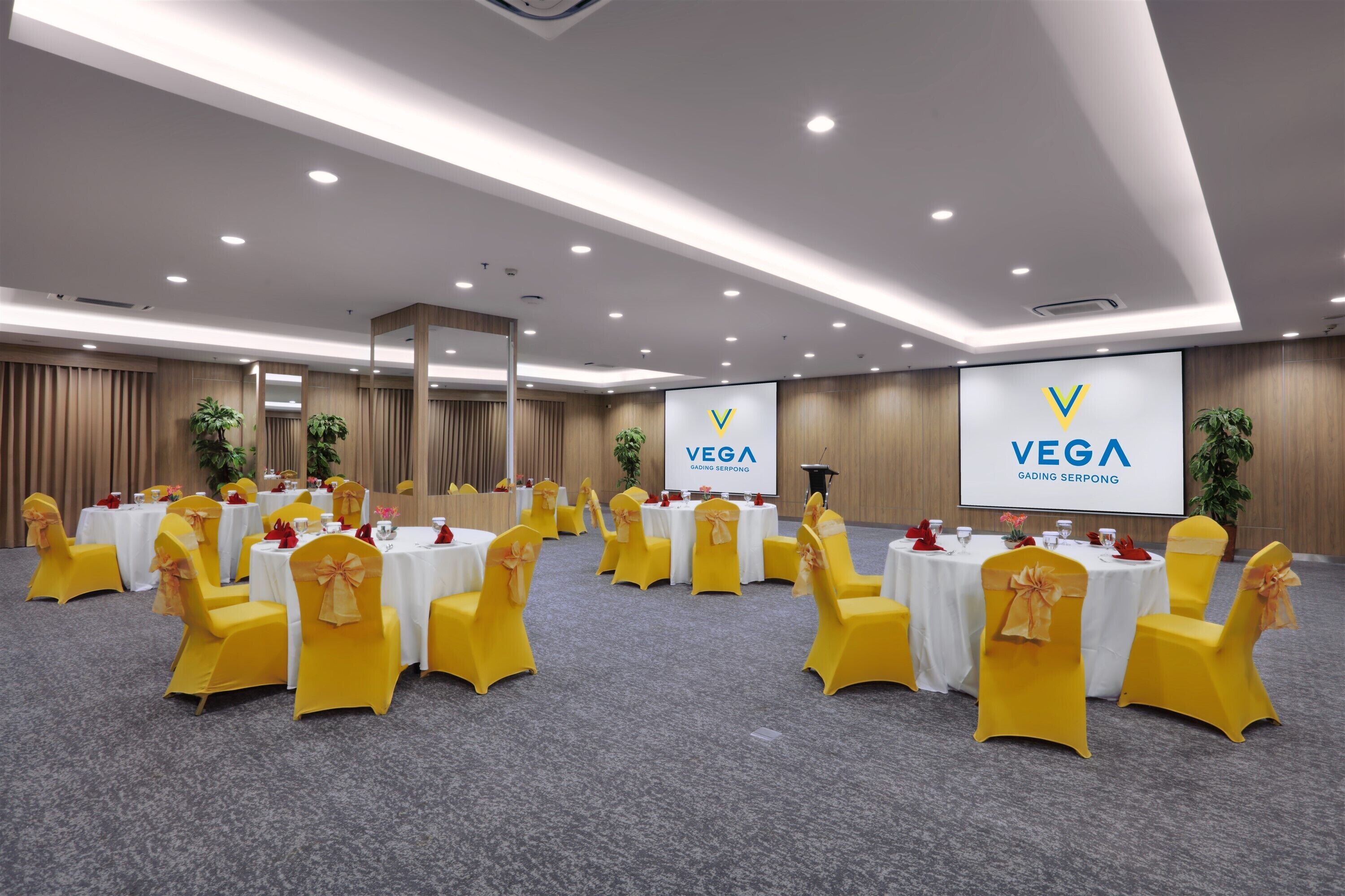 A conference room with round tables and yellow chairs, featuring a large screen displaying the logo 'VEGA GADING SERPONG'.