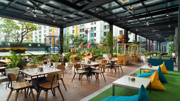 Un espace de restauration intérieur avec des tables et des chaises, entouré de plantes en pot et avec un terrain de jeux en arrière-plan.