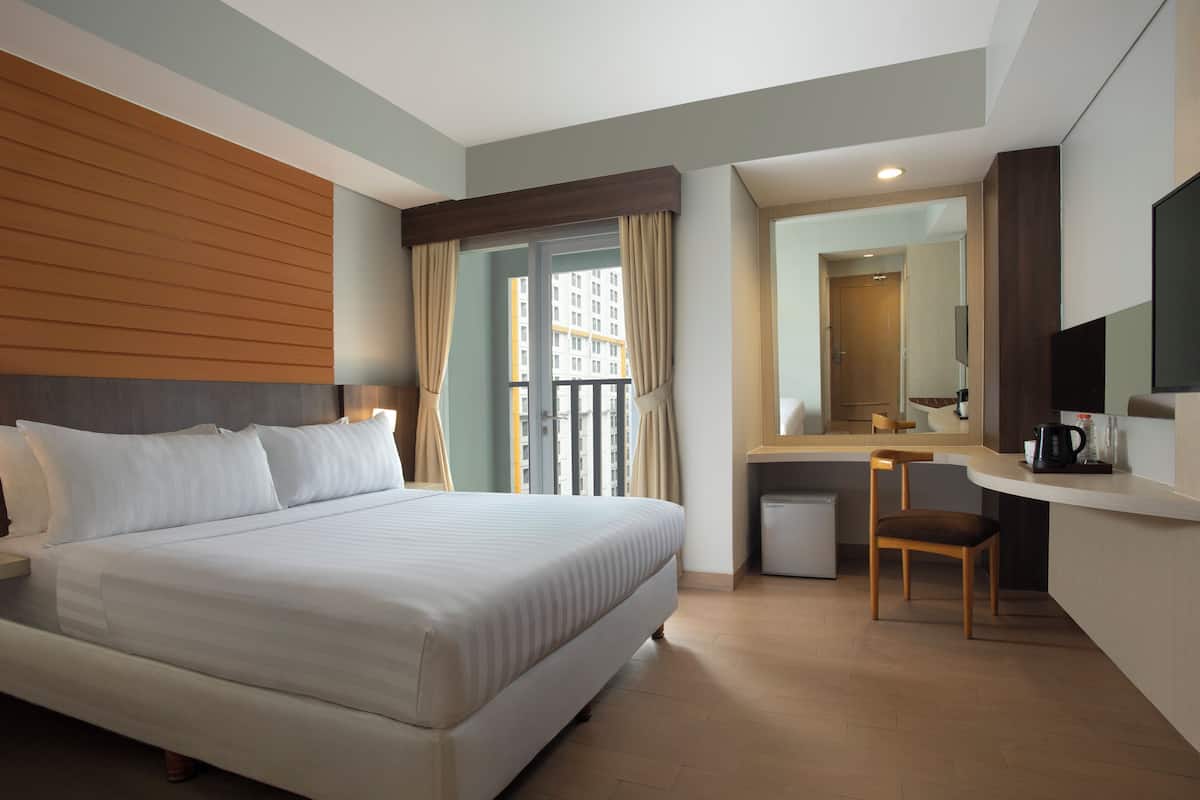 A hotel room with a large bed, a desk with a chair, a small kitchenette, and a balcony with curtains.