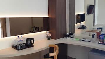 A hotel room with a curved desk, a chair, a coffee maker, water bottles, a TV, and a small shelf with snacks.