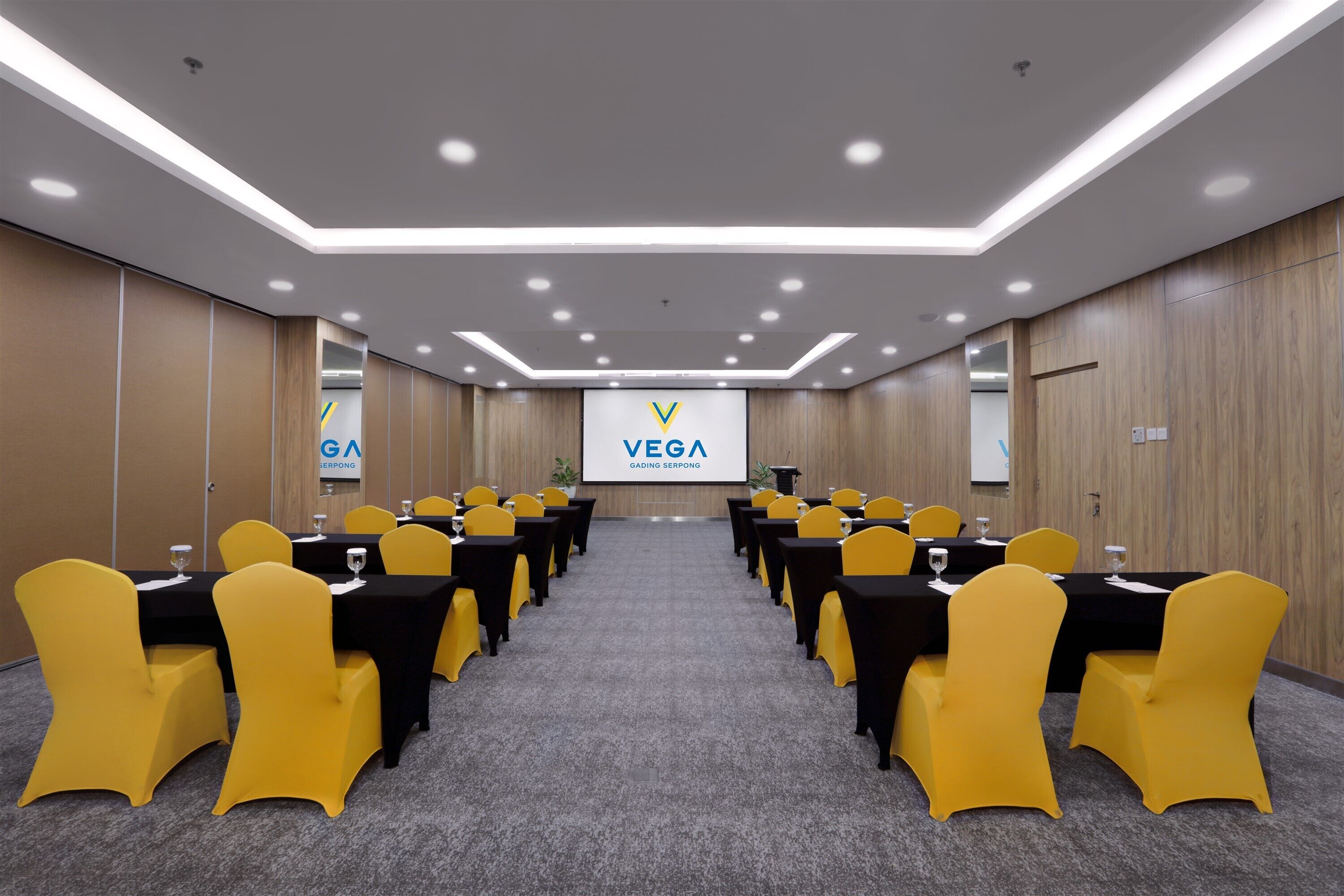 A conference room with tables and chairs, a large screen displaying 'VEGA GADING SERPONG', and a modern ceiling design.