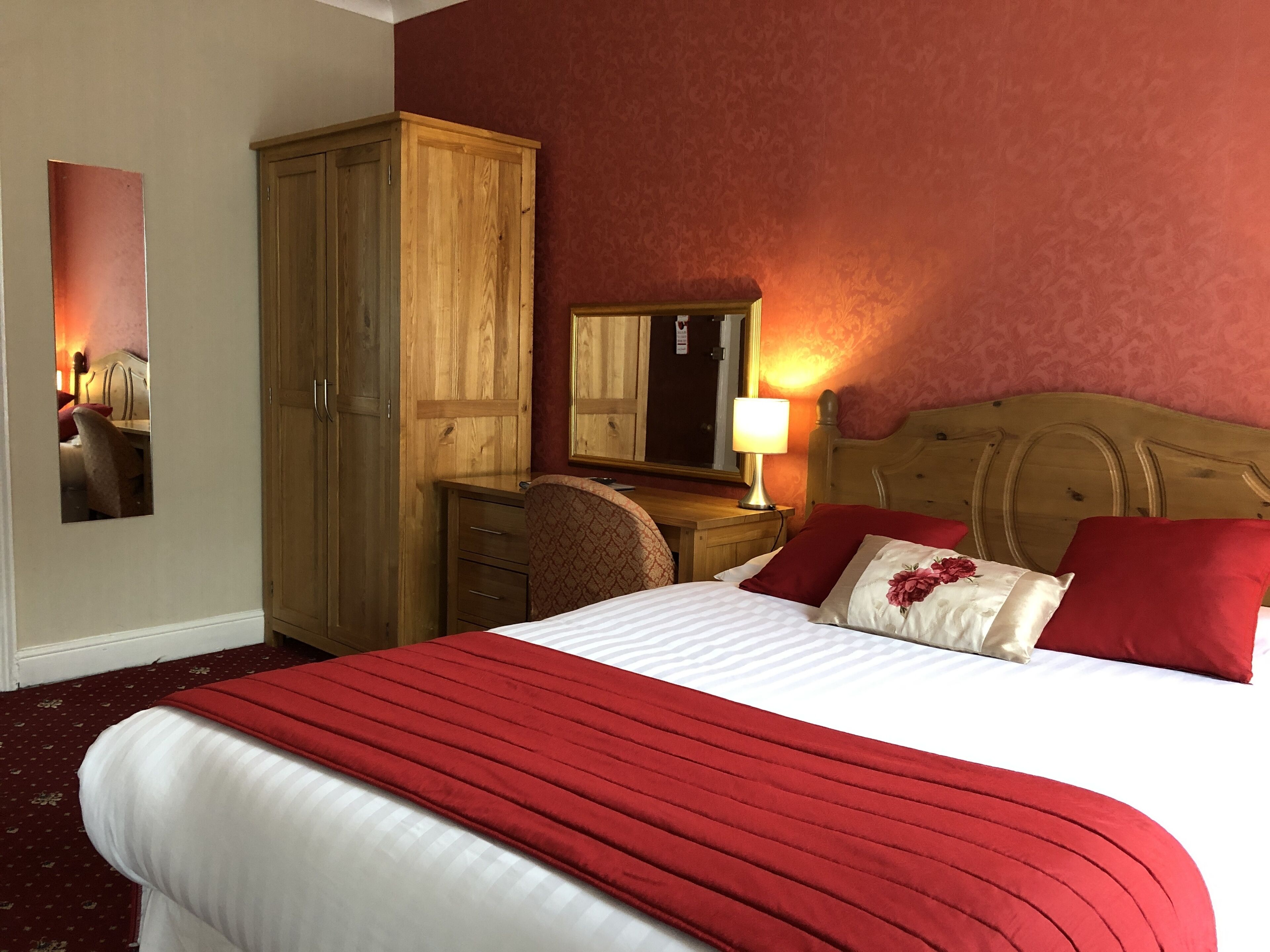 A hotel room with a large bed, a wooden wardrobe, a desk with a chair, and a mirror on the wall.