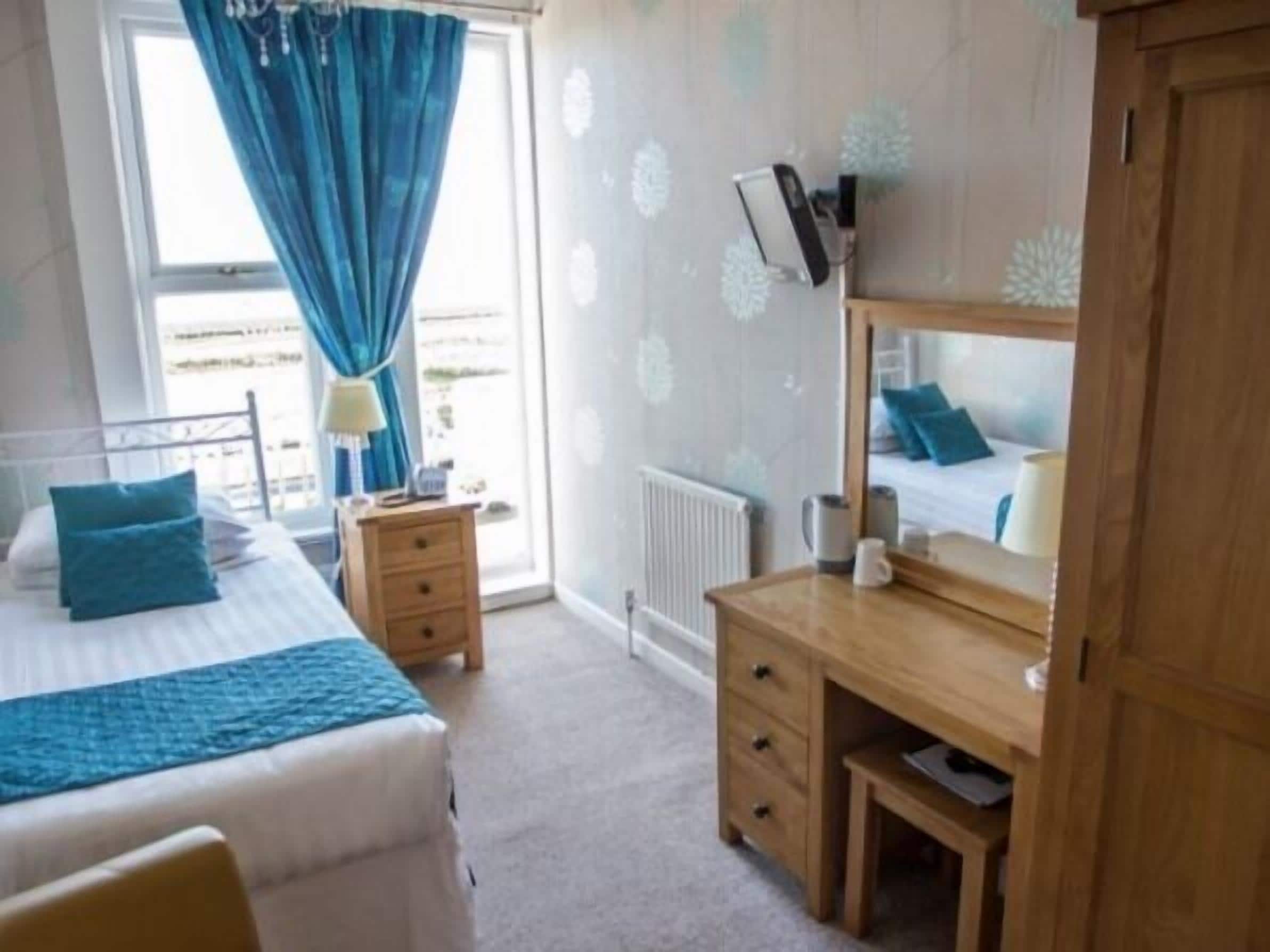 A bedroom with a bed, a wooden desk, a mirror, a radiator, and a window with curtains.