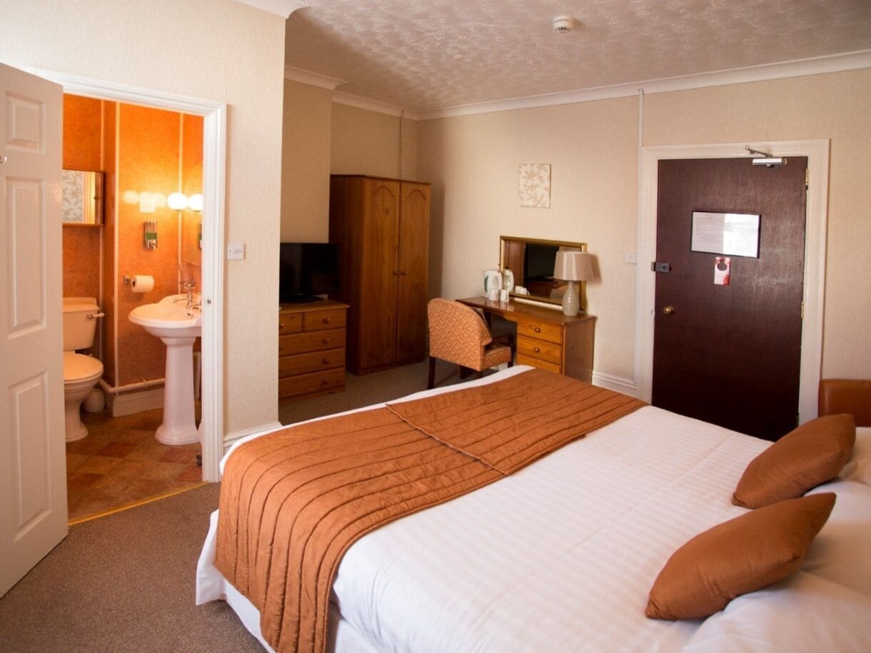 Quarto de hotel com uma cama grande, uma escrivaninha, uma cadeira, um guarda-roupa, banheiro com pia e vaso sanitário, e uma porta com um aviso.