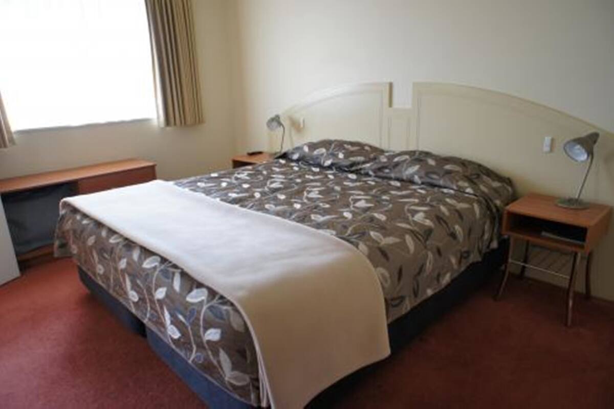 Ein Einzelbett mit gemustertem Bettbezug, ein Nachttisch mit Lampe und ein Fenster mit Vorhängen.