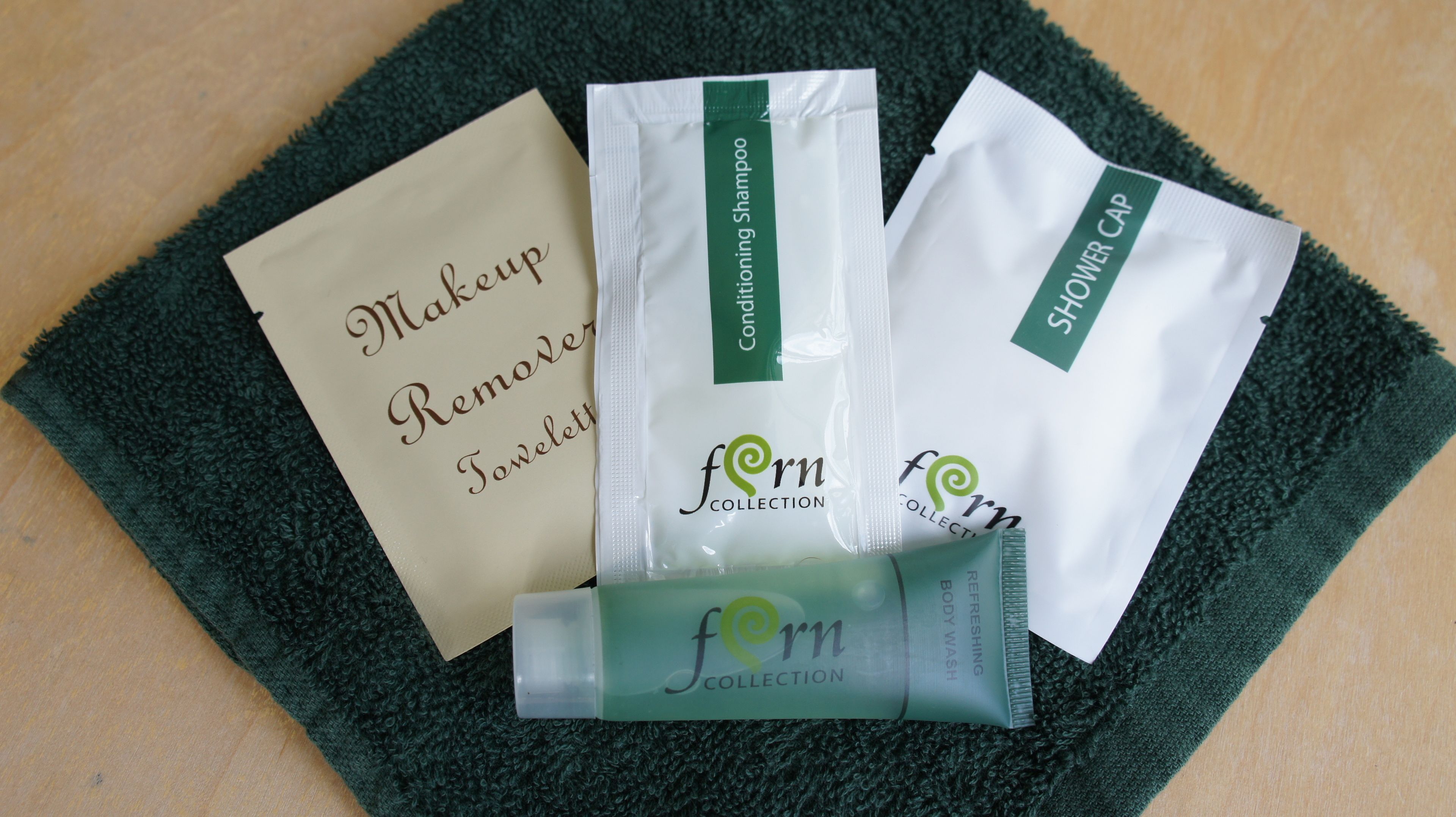 A package of makeup remover, a conditioning shampoo, a shower cap, and a body wash on a green towel.