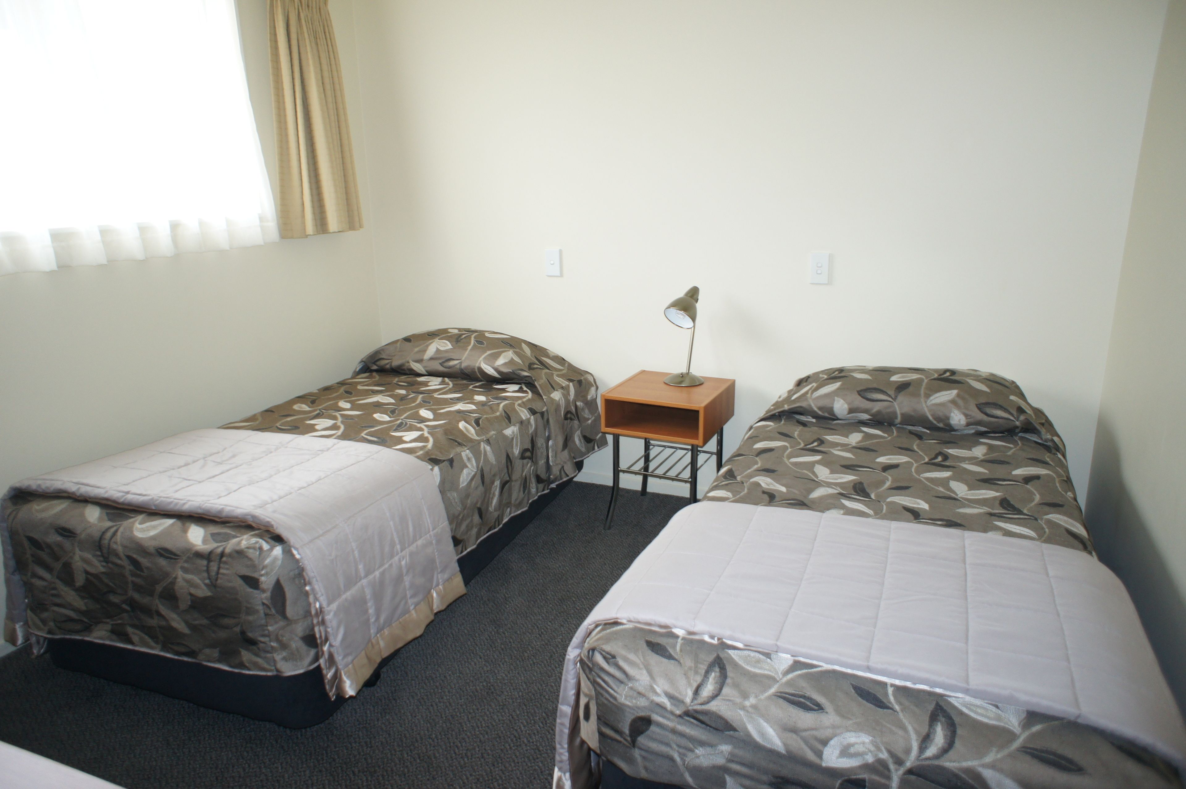 Una habitación con dos camas individuales, una mesita de noche con lámpara y una ventana con cortinas.