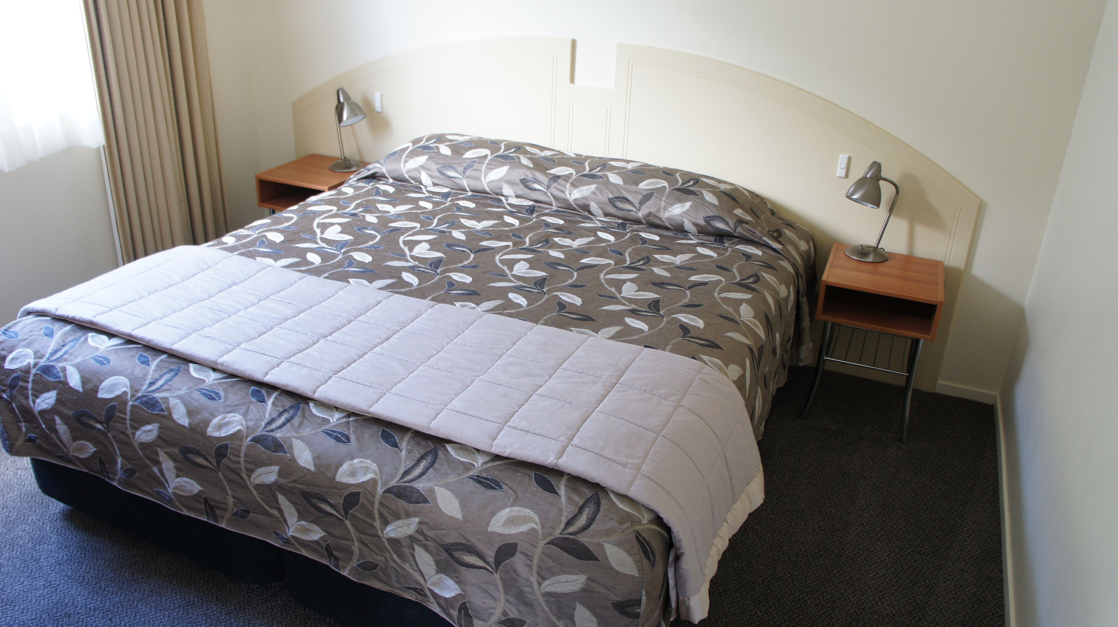 A neatly made bed with a floral-patterned duvet and pillows, two bedside tables with lamps, and a headboard with a light switch.