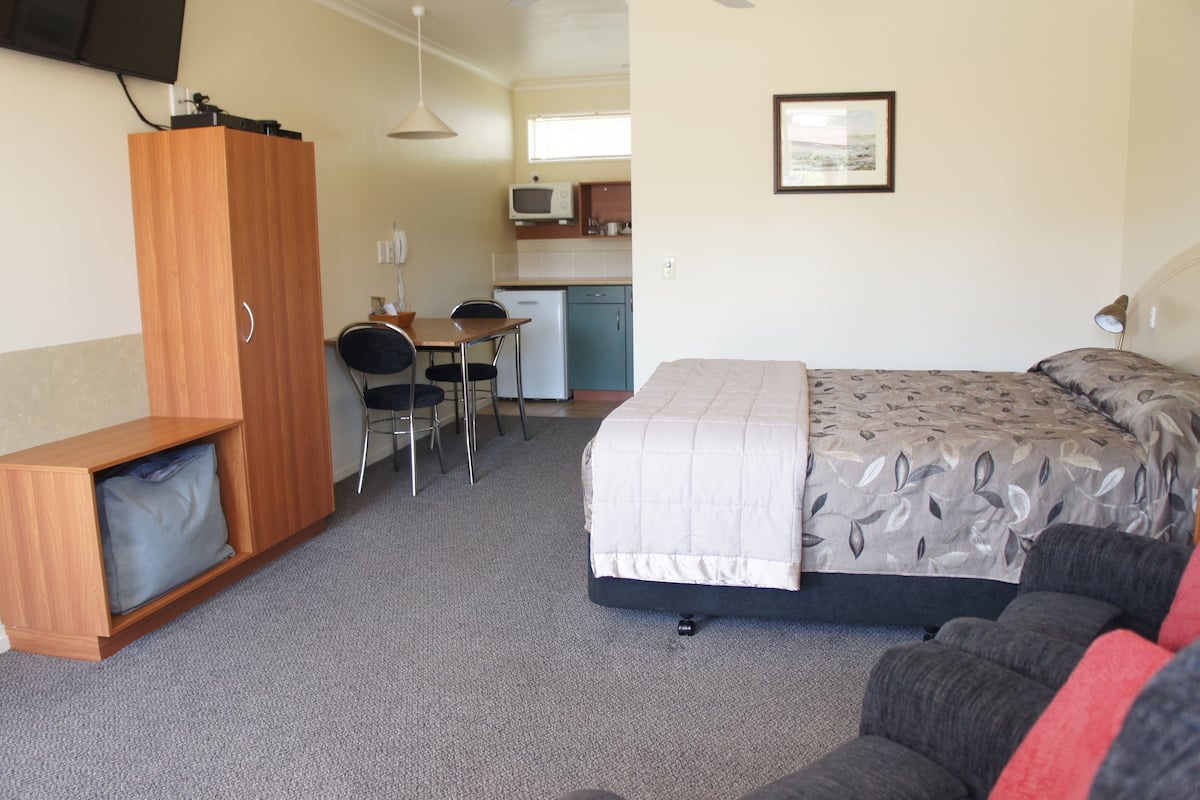Ein kleines Hotelzimmer mit Bett, Schreibtisch, Küchenzeile und an der Wand montiertem Fernseher.