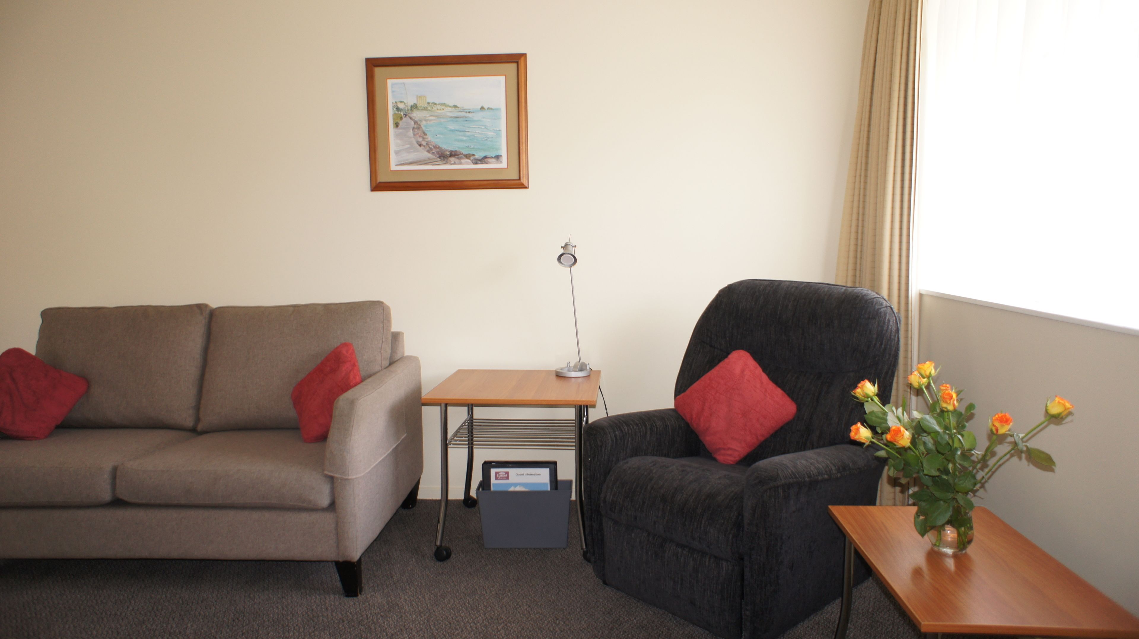 Une pièce avec un canapé gris, un fauteuil noir, une petite table en bois sur laquelle est posé un vase de fleurs, un tableau encadré au mur et une lampe sur une table d’appoint.