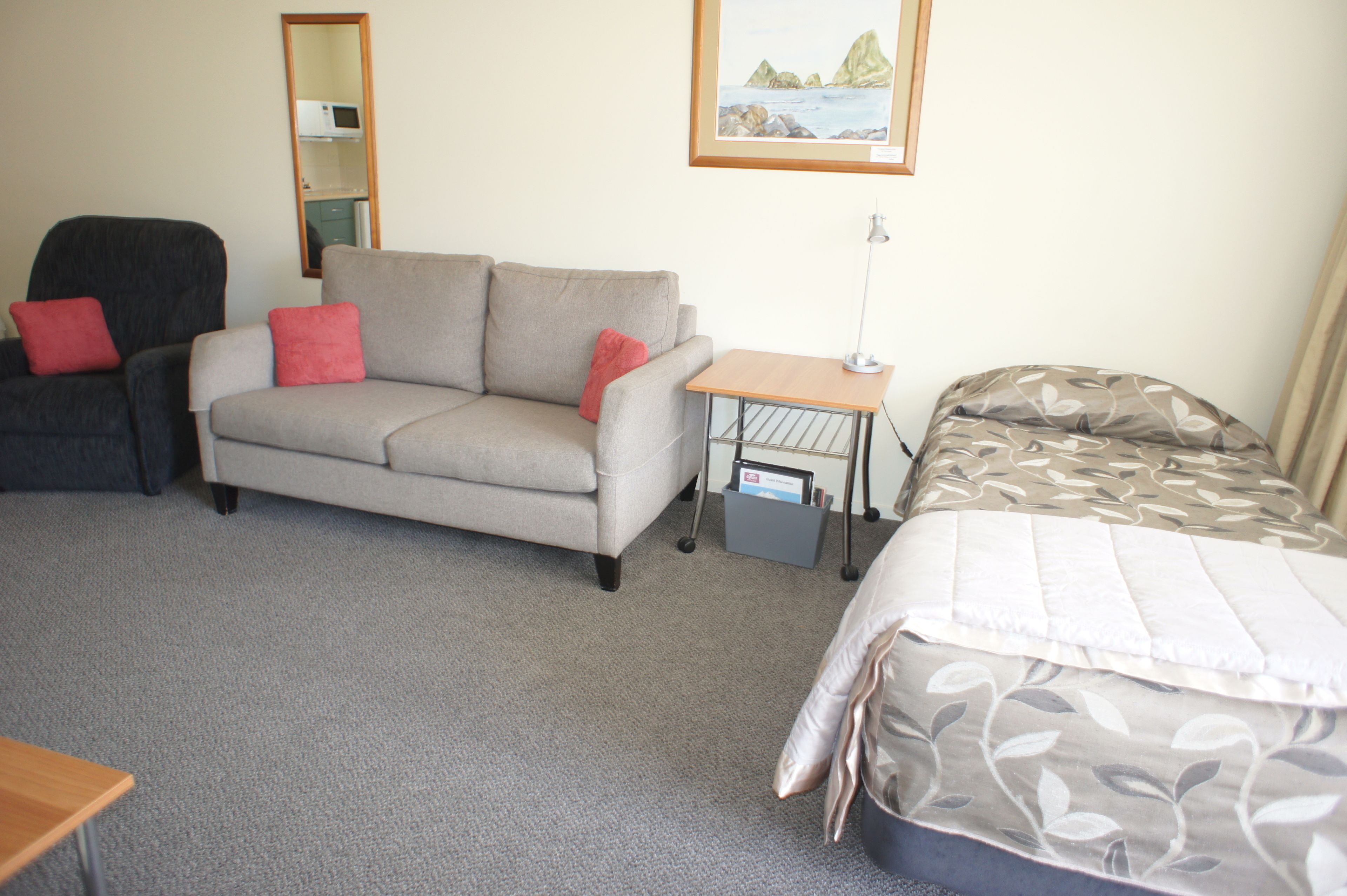 A hotel room with a grey sofa, a small table with a lamp, a bed with a patterned bedspread, and a mirror on the wall.