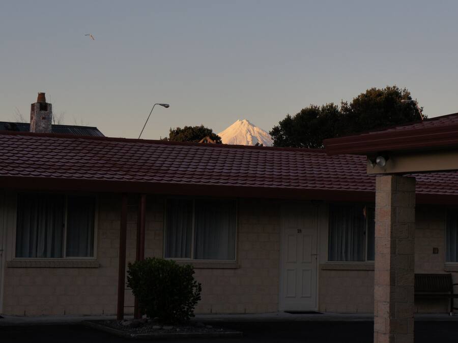 Un motel avec un toit recouvert de tuiles rouges, des chambres numérotées et une vue sur une montagne en arrière-plan.