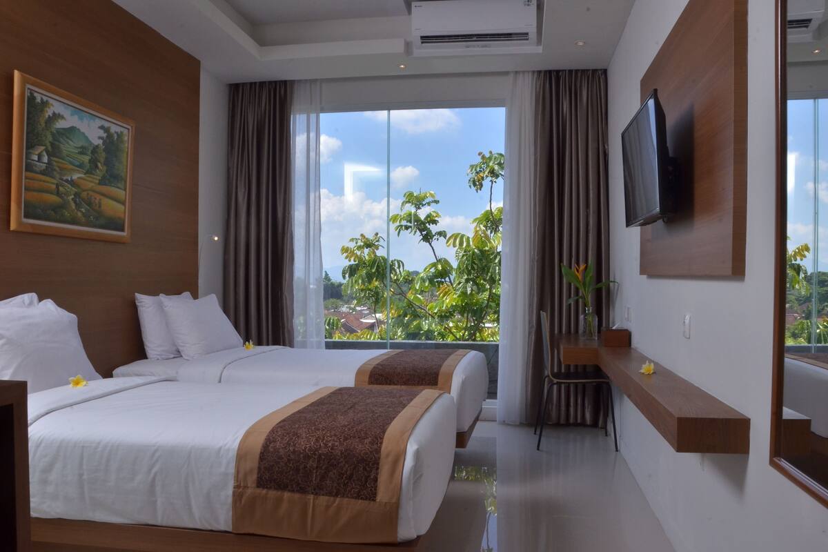A hotel room with two beds, a TV, a large window with a view, and a painting on the wall.
