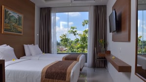 A hotel room with two beds, a TV mounted on the wall, and a large window offering a view of greenery and buildings.