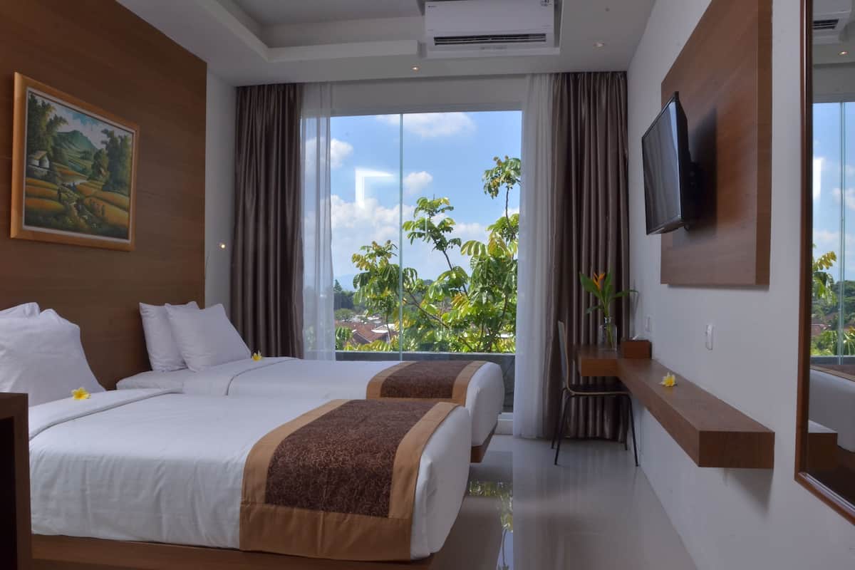 Une chambre d’hôtel avec deux lits, un téléviseur fixé au mur et une grande fenêtre donnant sur un paysage verdoyant et des bâtiments.