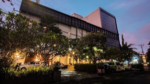A modern building with a flat roof and large windows, surrounded by greenery and parked cars.