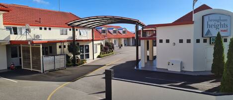 Entrada de un hotel con un paseo techado, un cartel que dice 'Brougham Heights Motel' y un camino de entrada con puerta de reja.