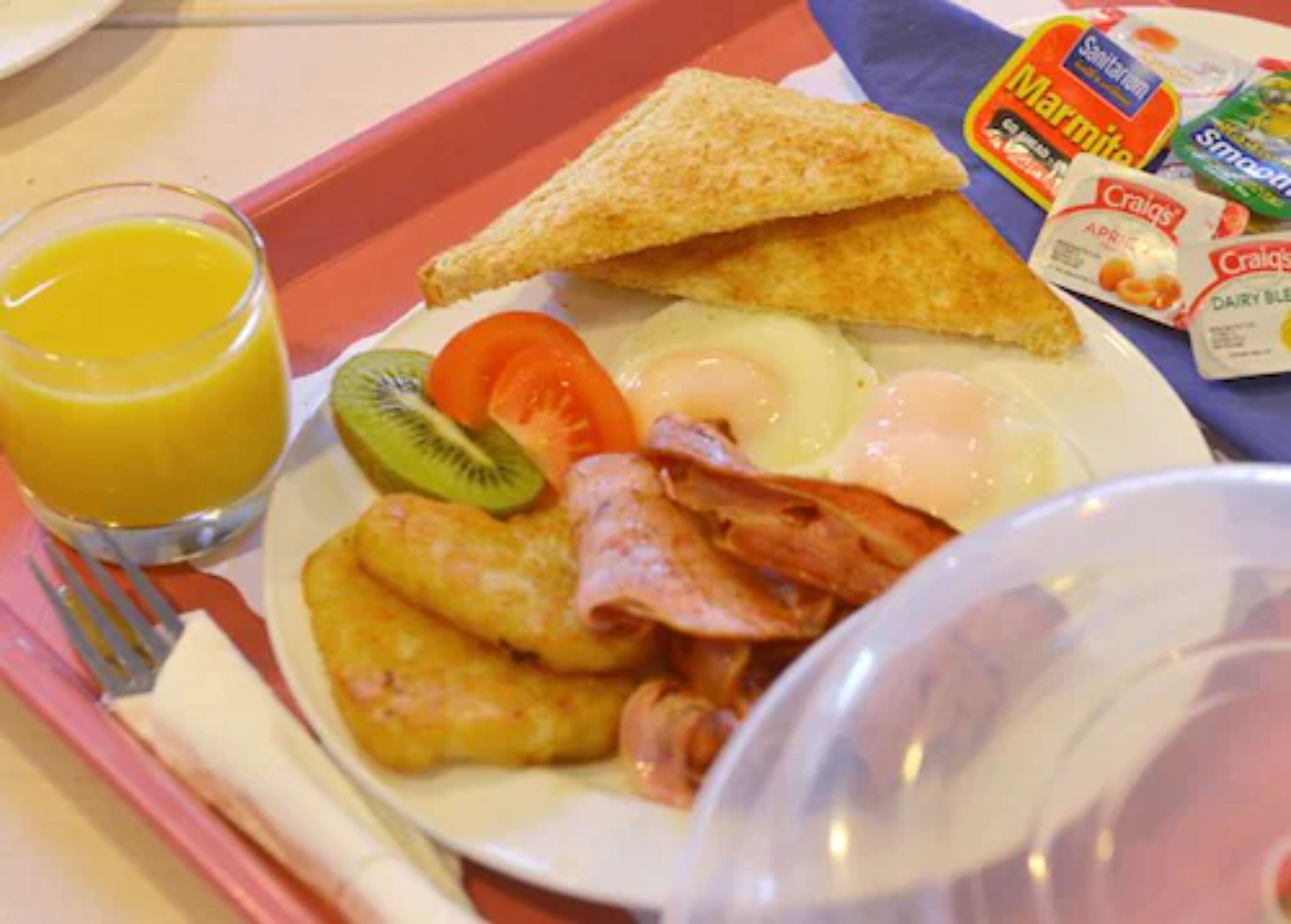 Een ontbijt bord met toast, eieren, spek en fruit, plus een glas sinaasappelsap en verpakte snacks.