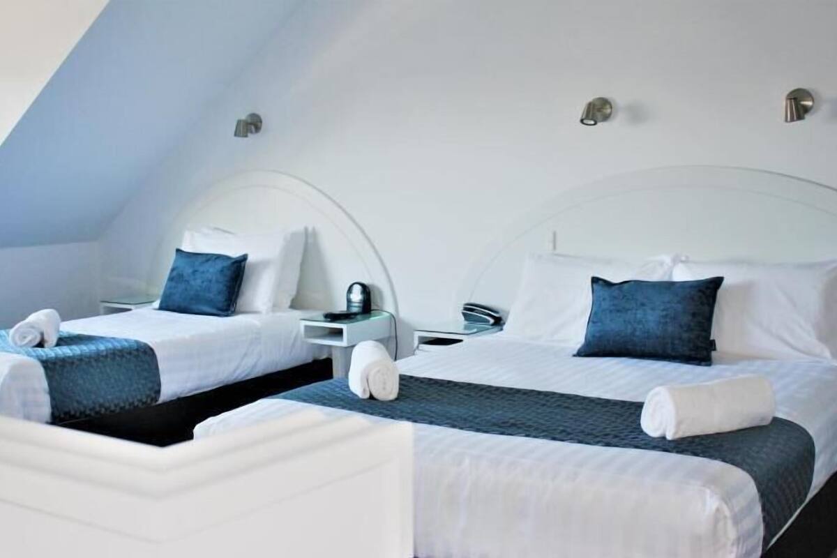 Twee eenpersoonsbedden met witte lakens, een klein nachtkastje met een lamp, en een gebogen plafond.