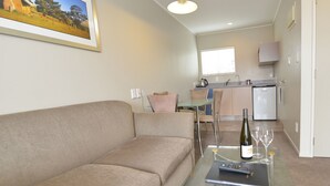 Una sala de estar con un sofá color crema, una mesa de centro de vidrio con un control remoto, una botella de vino y vasos. Se ve una cocina al fondo con un refrigerador y una ventana.