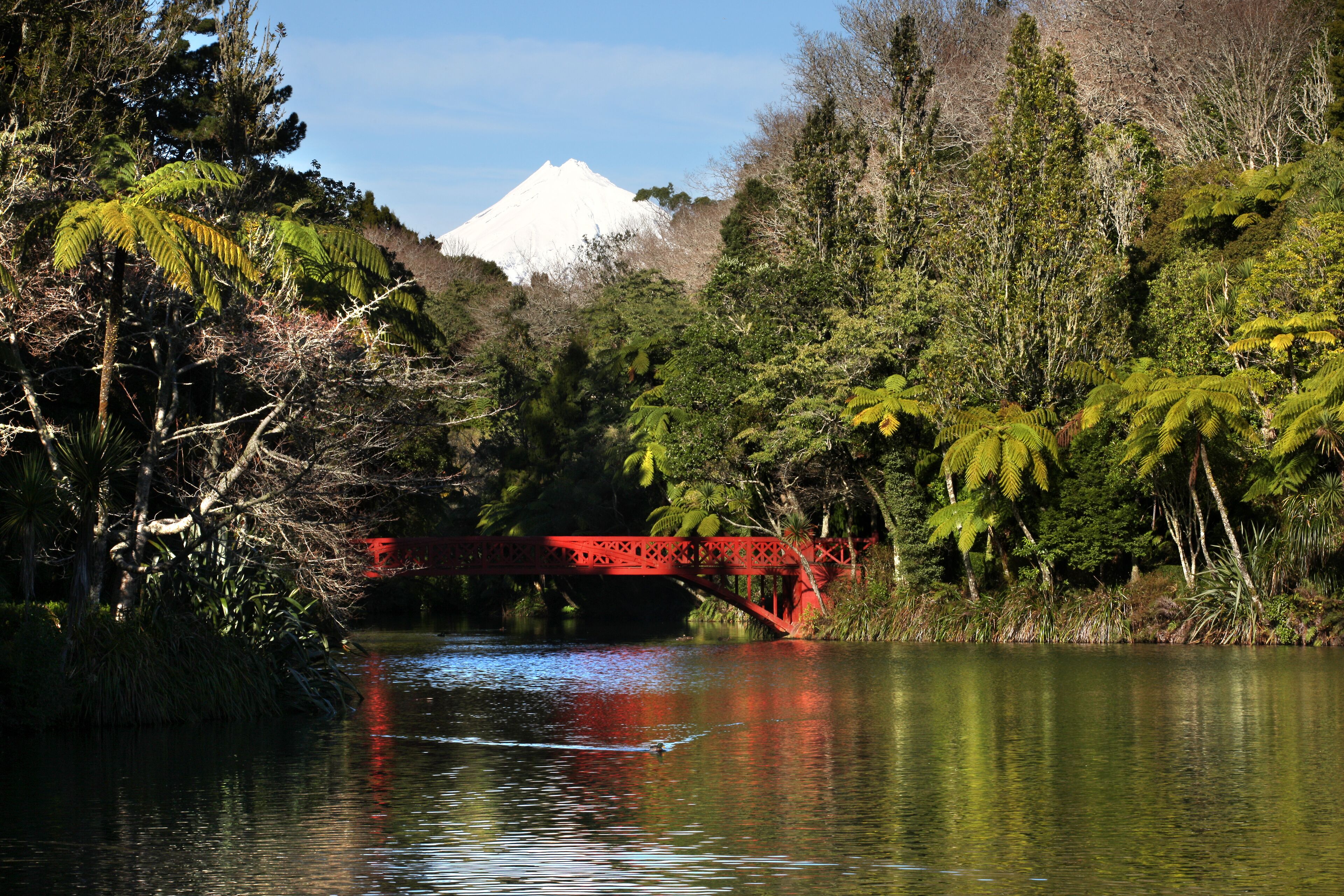 A red bridge over a calm river, surrounded by dense greenery and a snow-capped mountain in the background.