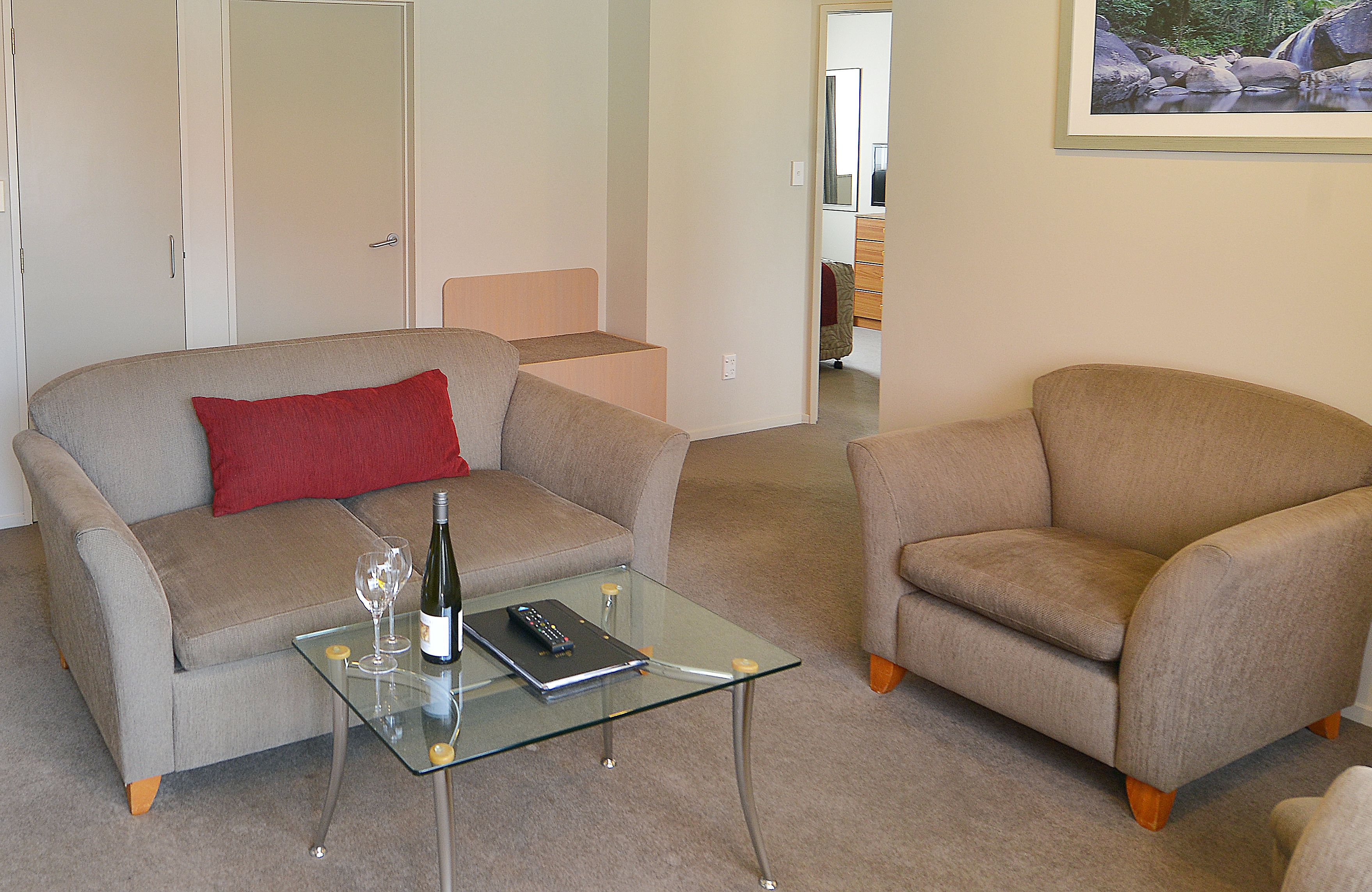 Un salon avec un canapé, un fauteuil, une table basse en verre et un coussin rouge.