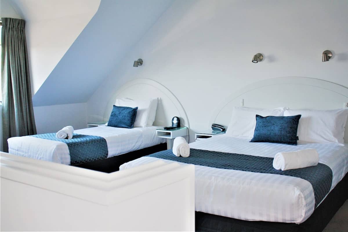 Twee eenpersoonsbedden met witte hoofdborden en blauw beddengoed, een nachtkastje met een lamp en een raam met gordijnen.