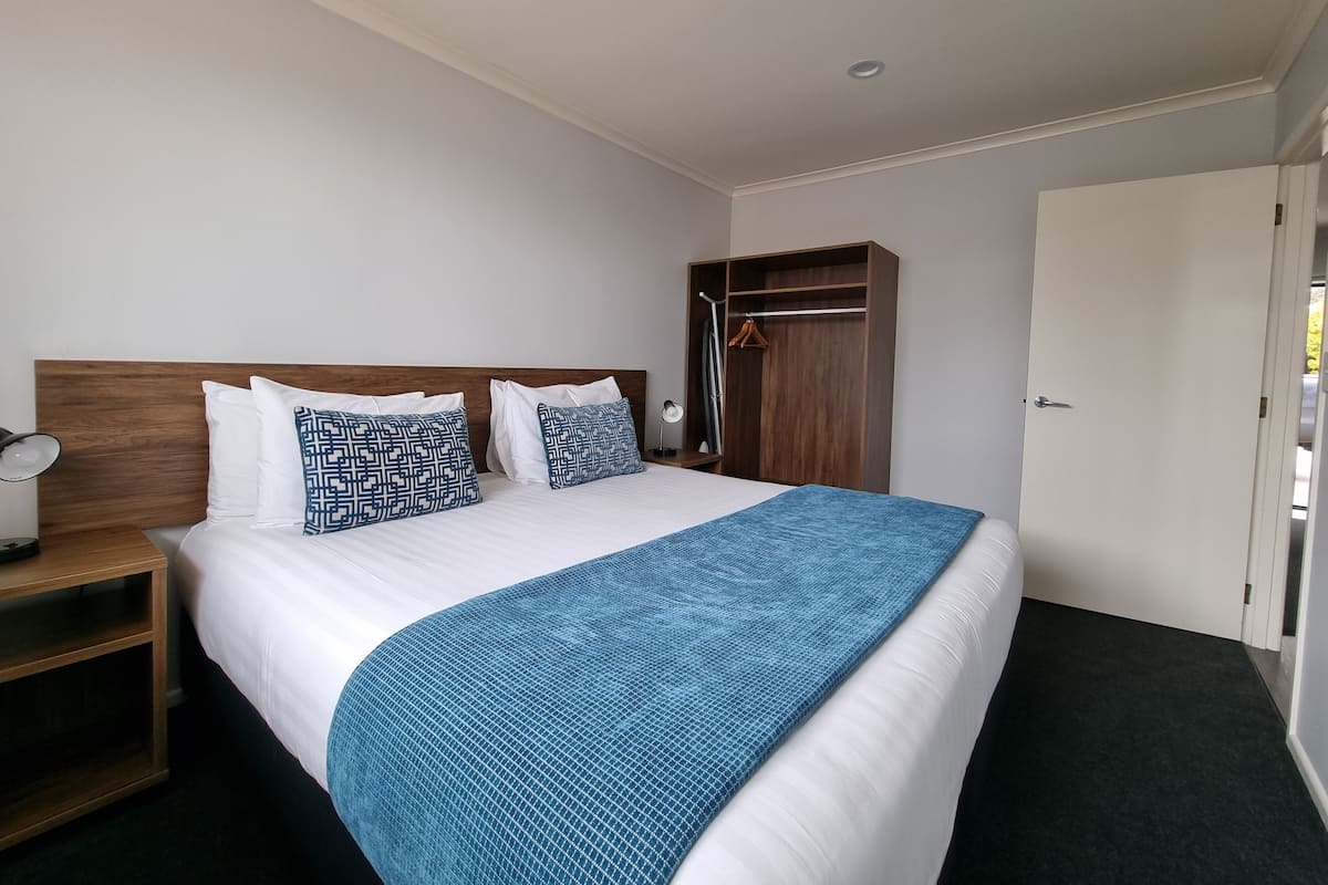 Hotelkamer met een houten hoofdbord, een bed met kussens in blauw en witte patronen, een blauwe gebreide deken, een nachtkastje met een lamp en een garderobe met spiegel.