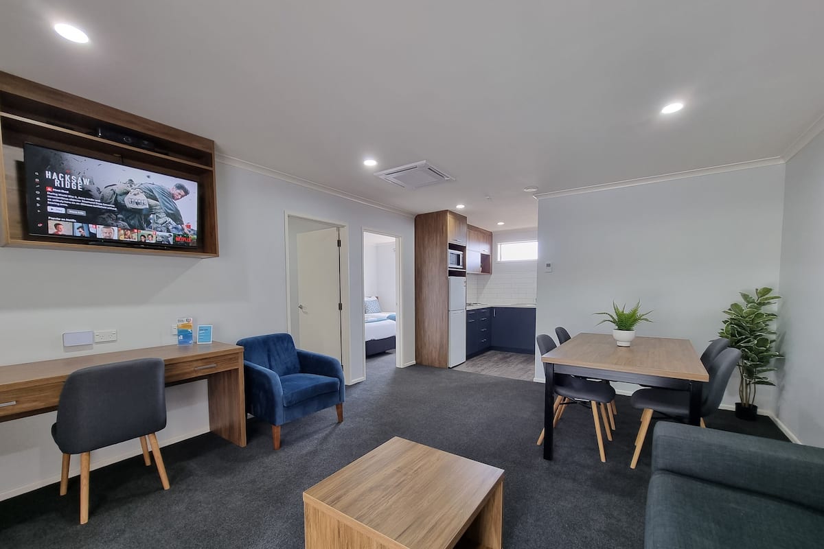 Een moderne hotelkamer met een houten bureau, een blauwe fauteuil, een houten salontafel, een eettafel met stoelen, een keukenhoek en een aan de muur bevestigde televisie.