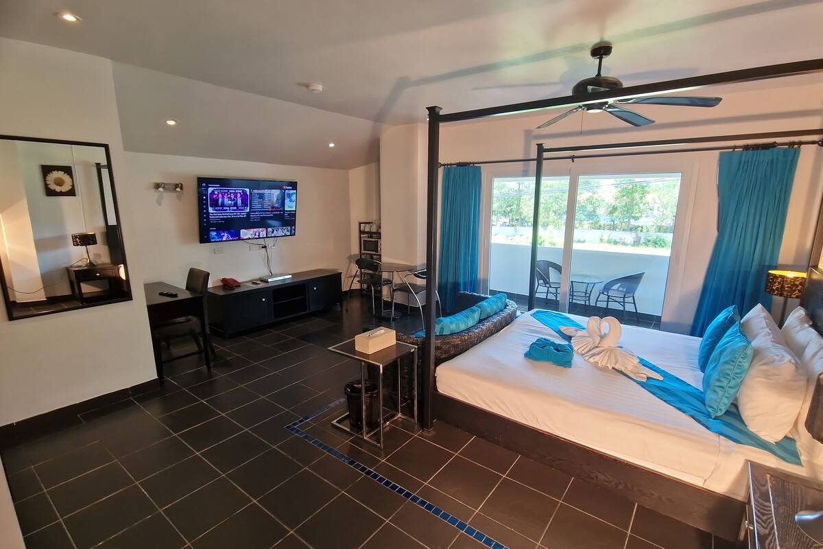 Habitación de hotel moderna con una cama grande, un televisor, un balcón con sillas y un ventilador de techo.