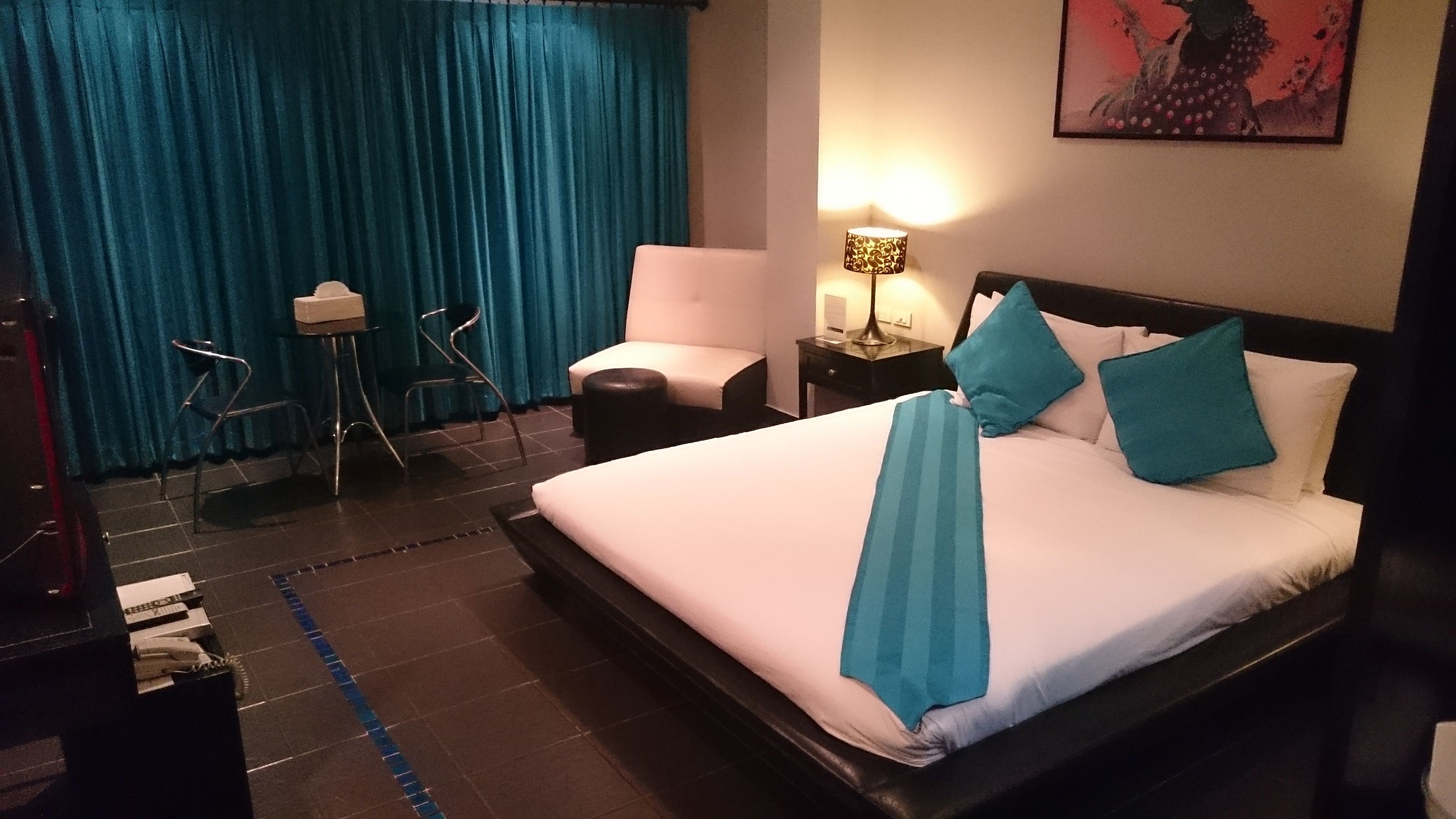 A hotel room with a large bed, a desk with a lamp, a chair, and a blue curtain.