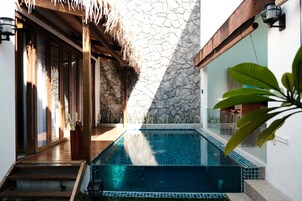 A swimming pool with a stone wall and wooden deck, surrounded by glass doors and a thatched roof.