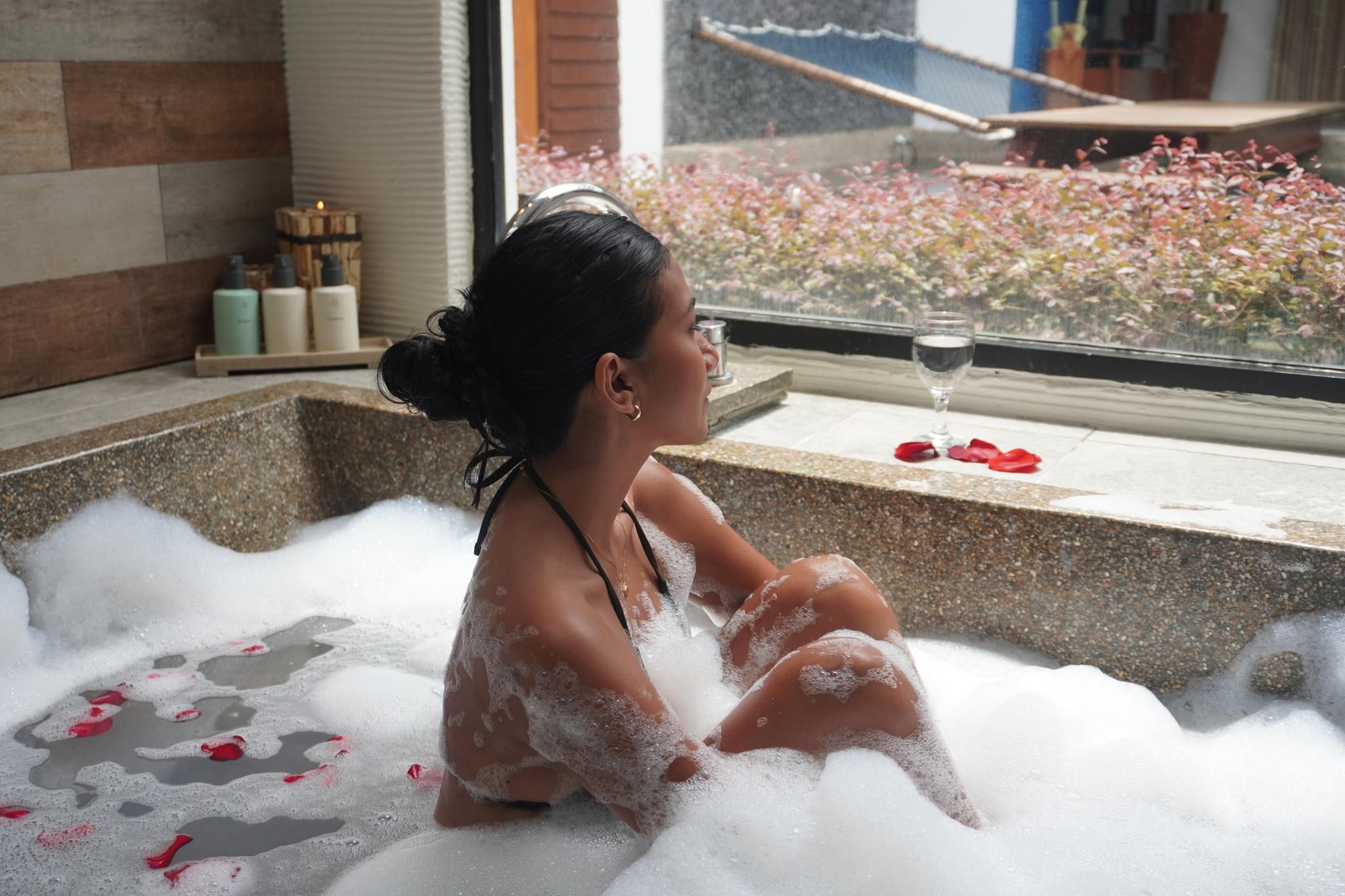 Una persona sentada en una bañera llena de burbujas, con pétalos de rosa esparcidos alrededor.