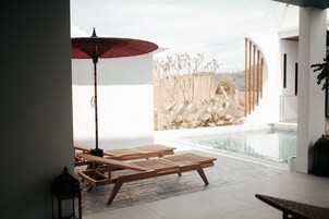 Zona junto a la piscina con sillones de descanso y un paraguas rojo, con vistas a una piscina y un muro de piedra.