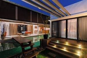A modern outdoor pool area with a wooden deck, a seating area, and a staircase leading to a covered patio.