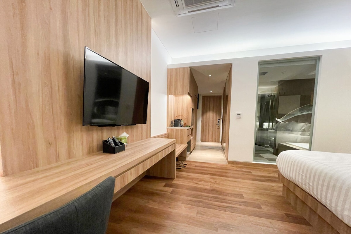 A modern hotel room with a wooden floor, a flat-screen TV mounted on the wall, a desk with a chair, a bed, and a glass door leading to a bathroom.