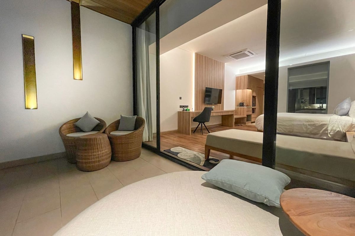 A modern hotel room with a large bed, two wicker chairs, a desk, and a television.